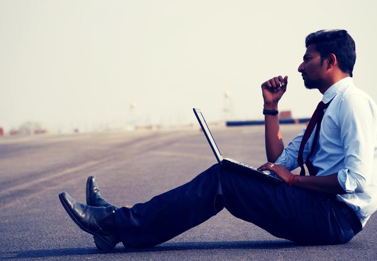 člověk v modré košili s kravatou sedí na silnici a má na klíně laptop