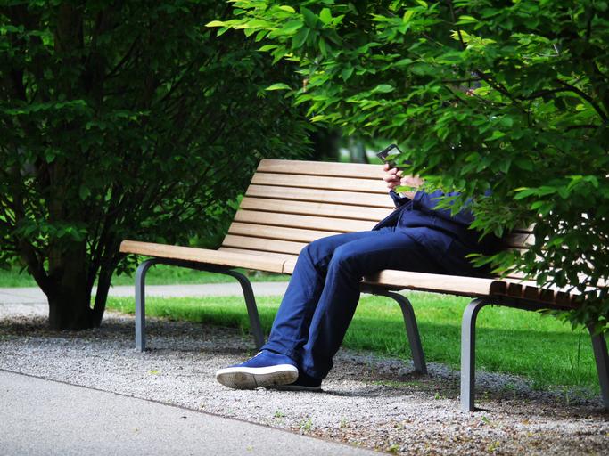 park, lavička, na ní sedí muž v modrém obleku s mobilem v ruce, tvář a půl těla má za stromem